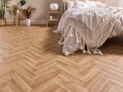 Bedroom with light herringbone wood floor, beige bedding, a chunky tasselled throw, and a small bedside table with a lamp and potted plants.